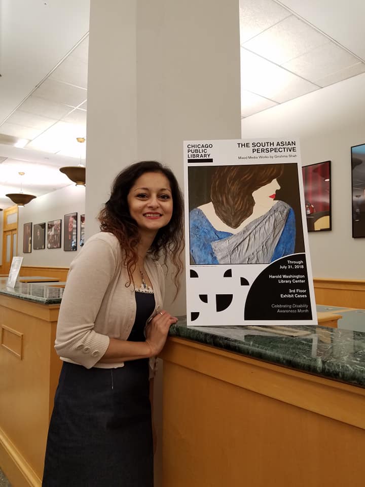 Grishma, A South Asian woman of color with brown curly hair, wearing a white sweater, blue frock and red lipstick smiles at the camera. She stands next to her exhibit sign that says, "The South Asian Perspective" at the Harold Washington Library.