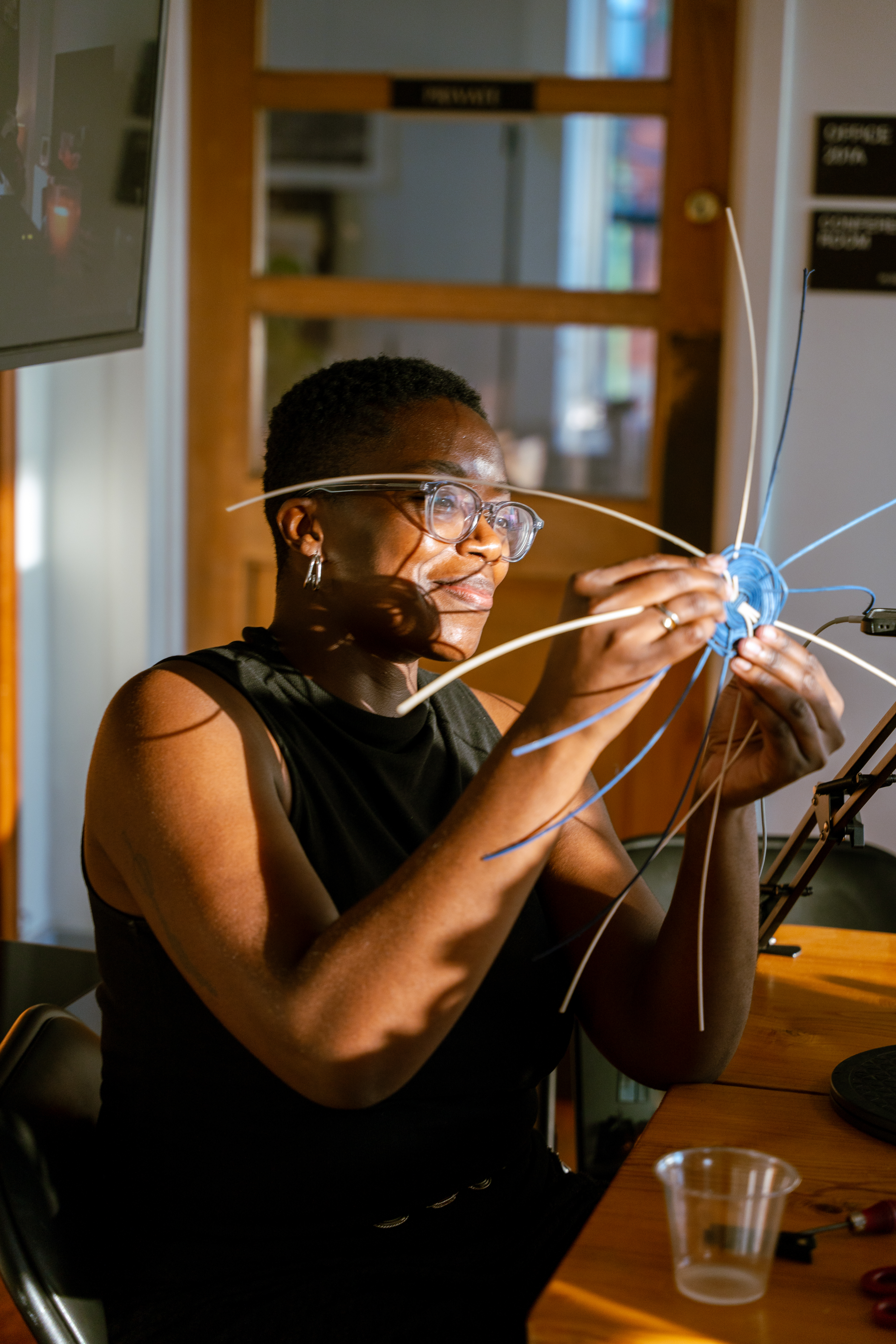 A black woman with glasses, queer presenting with short black hair holds basket weaving materials in the sunlight.