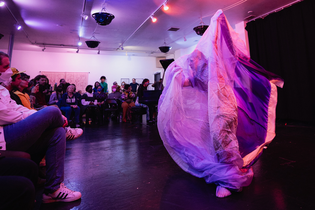 Sara Zalek performing at Freedom From and Freedom To. The audience can be seen in a u shape and Sara is seen through a luminescent fabric. 2024