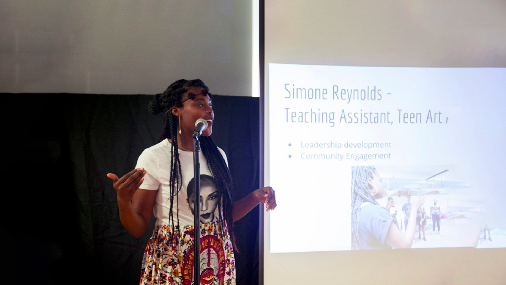 Simone, a tall dark-skinned person, wearing a Sade graphic t-shirt and multi-colored Ankara skirt, is standing at a mic in front of a presentation projected onto the screen. She is describing her work as an intern at Arts + Public Life.