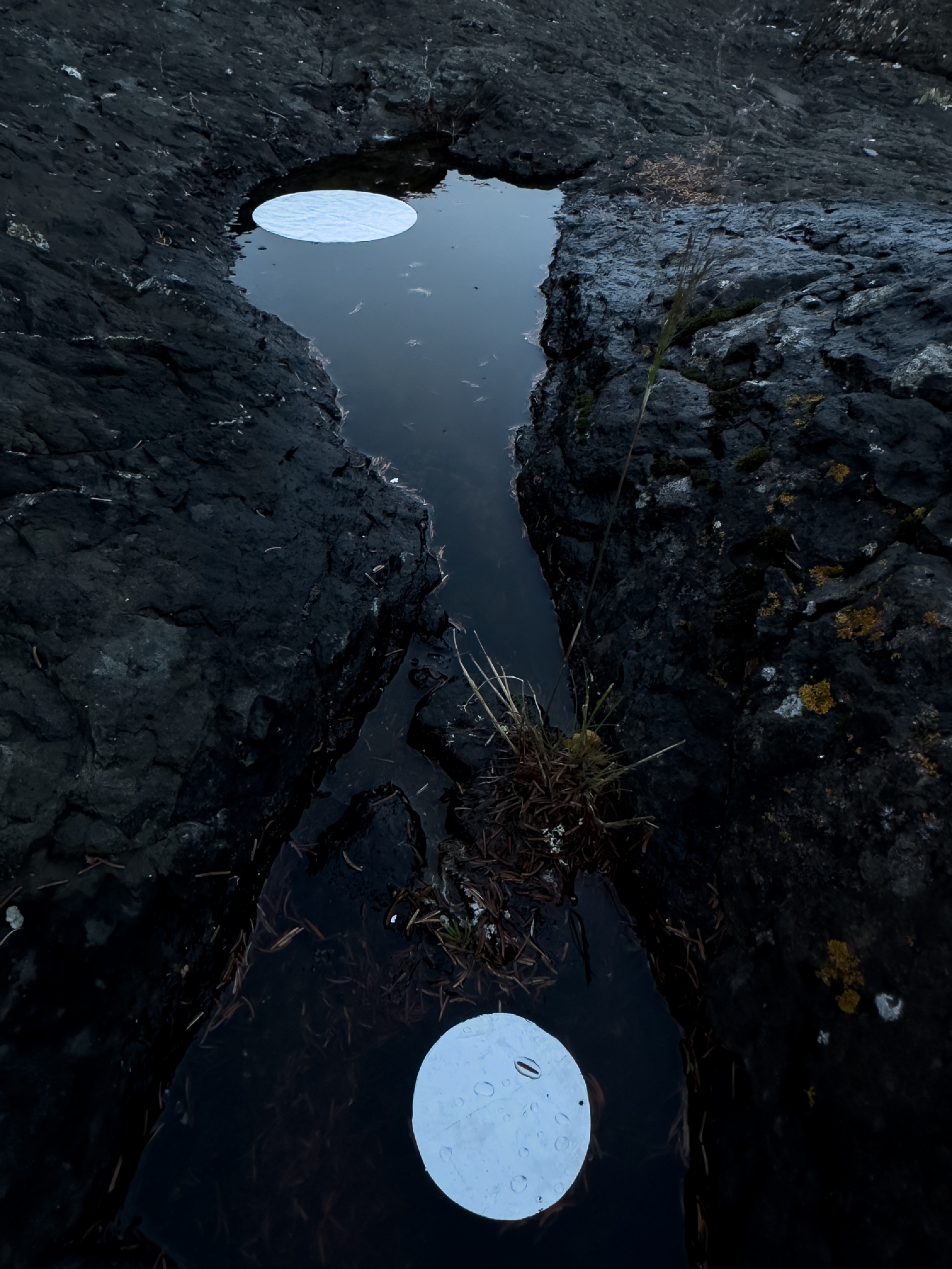 A small, still pool of water is surrounded by dark, jagged rocks. The smooth surface of the pool seems to reflect the sky, showing two bright, white circular shapes resembling moons or lights against the dark rock.
