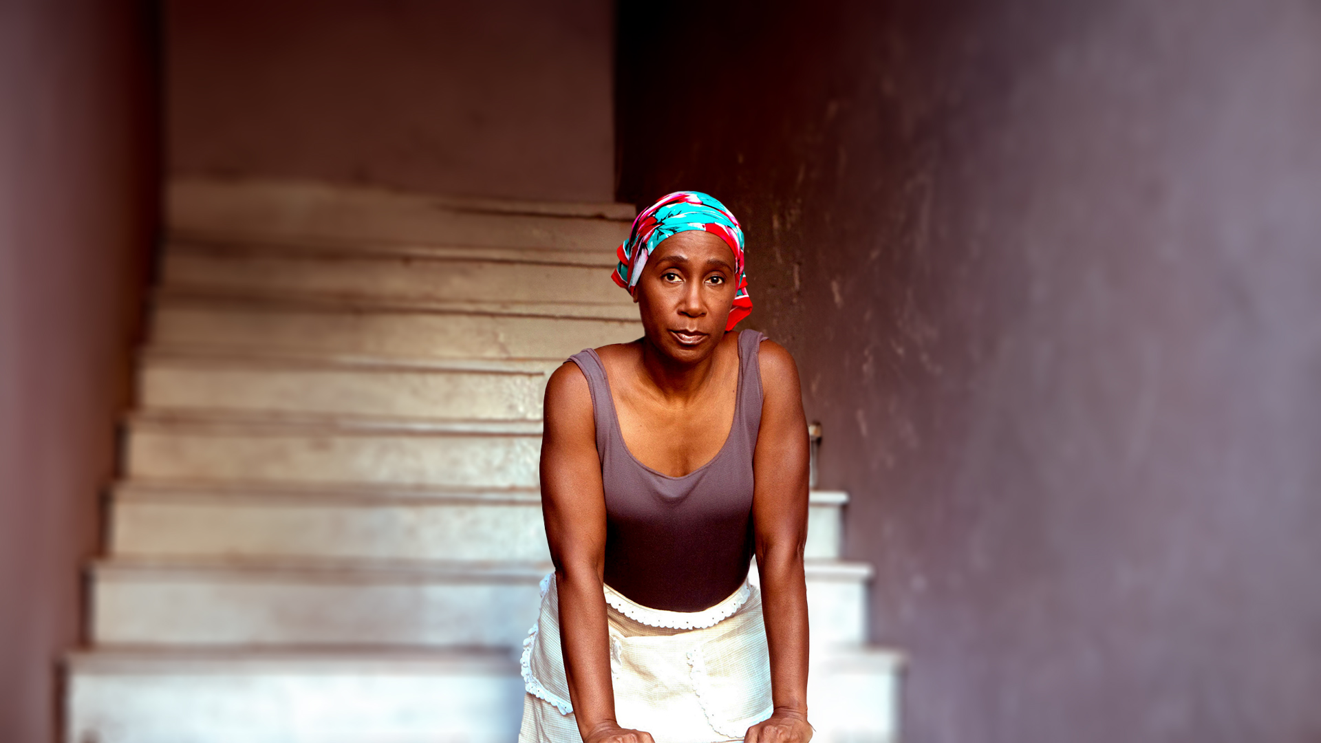 woman in hair scarf, brown tank, and white skirt looks deeply into the camera