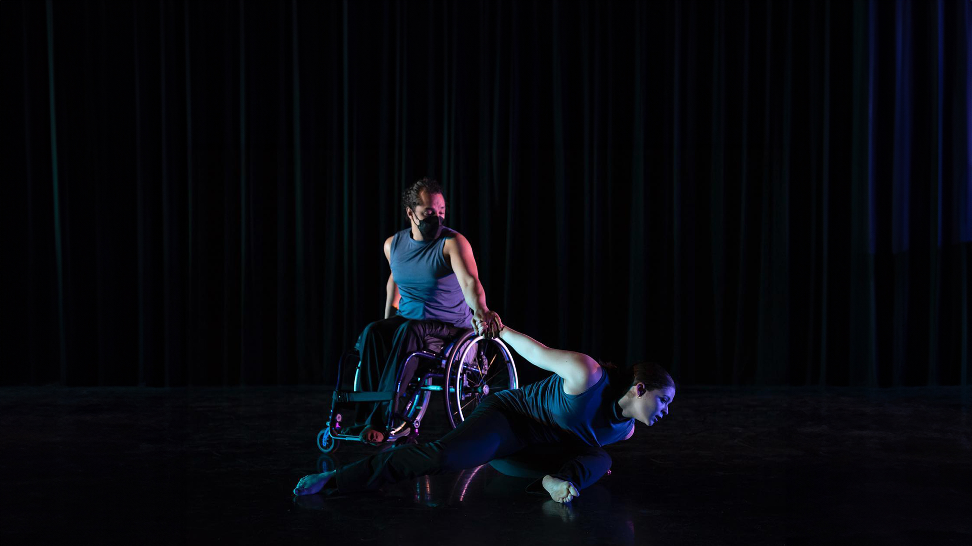 person in wheelchair holds person as they dance on dark stage