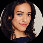 A headshot of a woman with medium brown skin and shoulder-length, softly layered dark brown hair styled in loose curls. She has warm-toned natural makeup, and wears a soft rose lipstick. She is wearing a navy blue top with a distinctive cut-out neckline,