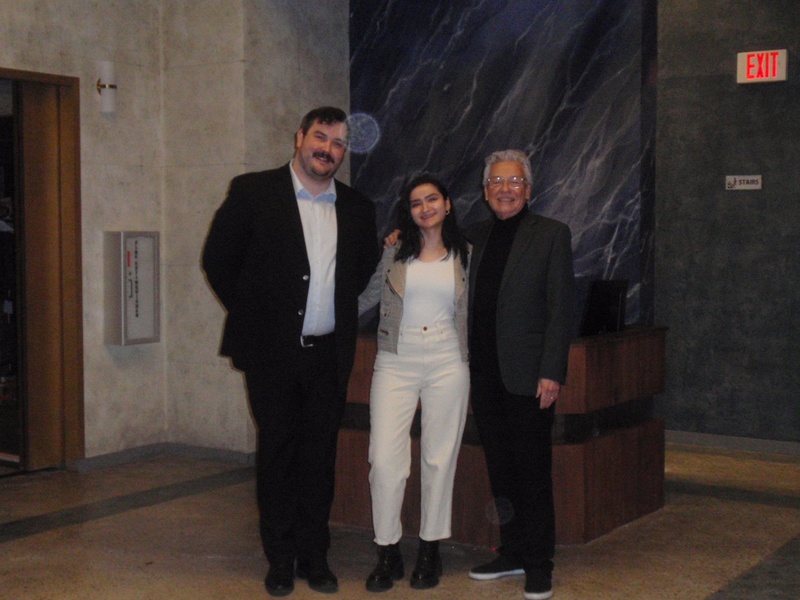  Three figures stand in a building lobby. They are smiling and staring at the camera. They are each wearing formal outfits with two of the wearing suit jackets/ 