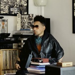 Black man short hair on the sides, long on the top, wrapped in black leather shawl, with sunglasses sitting in front of record shelves.
