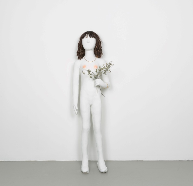  Photo of a sculpture in a white wall gallery. The sculpture is of a girl but has no features. The skin is white and it has brown hair, a gold necklace and is holding flowers. 
