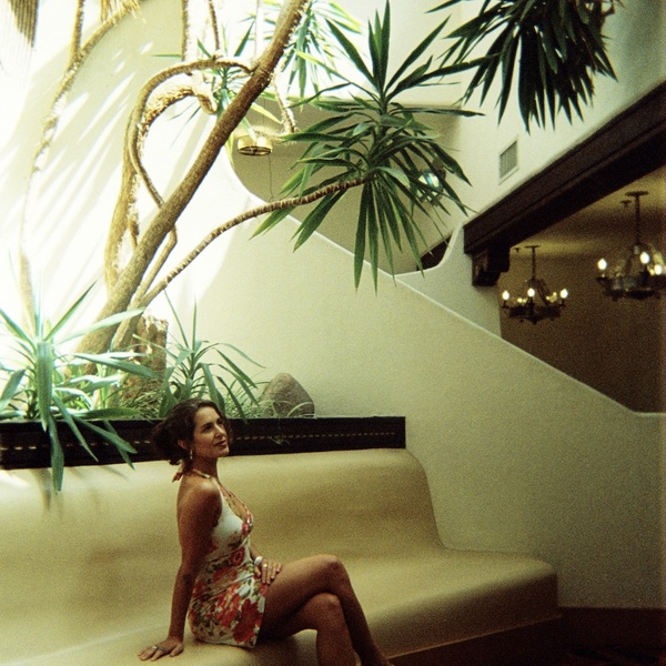  A femme-presenting person with long curly dark hair tied back poses on a yellow/beige chair with a large tree behind them. 
