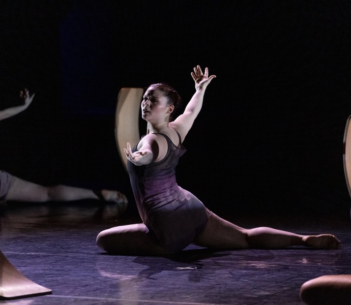  A white woman with red hair tied back performs on a dark stage. She is wearing a black leotard and is posed with both of her hands towards the ceiling. 