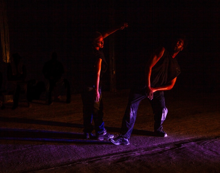  Two performers dance outside on a concrete floor. It is dark outside with reddish pink lighting. 