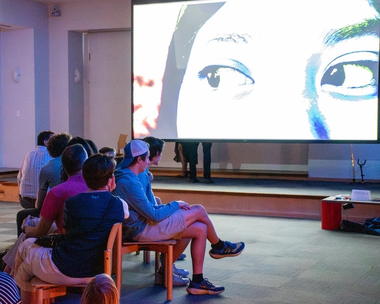  An audience sits and watching a projection on a video screen. The video is a close up on a pair of dark eyes. 