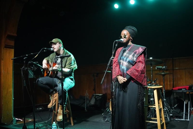 A Black Woman in a Hijab stands at a microphone. She is wearing sunglasses and a red shirt with a black skirt. A person is next to her sitting on a stool playing the guitar. 