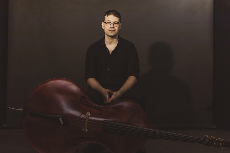  A white man with glasses and short dark hair looks at the camera. There is a dark backdrop and below the man a bass is resting on the floor. 
