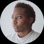 A Black woman standing in a room looking off to the side. She has a short haircut and is wearing a grey t-shirt, studded earrings, and a wavy peace sign chain. Part of a leaf from a monstera plant also stands behind her.