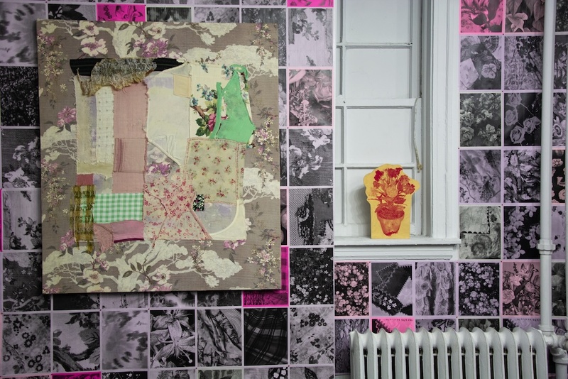  Image of a collaged fabric painting hanging on a wall next to a small image of red flowers printed on yellow paper. The wall behind the fabric painting is pint and has a grid of black and white images printed on it. 