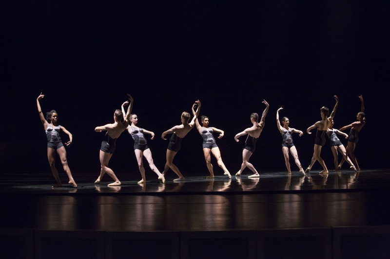  A group of ten dancers performer in an all black room. They are wearing matching outfits that are light grey. They have formed a line and are mirroring each other, their arm reaching up and their leg stepping back., 