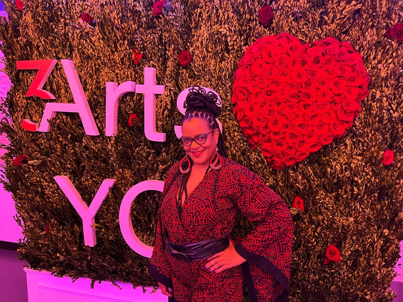  Black woman wearing red and black African print  jumpsuit in front of 3arts background and big red heart made of red flowers 