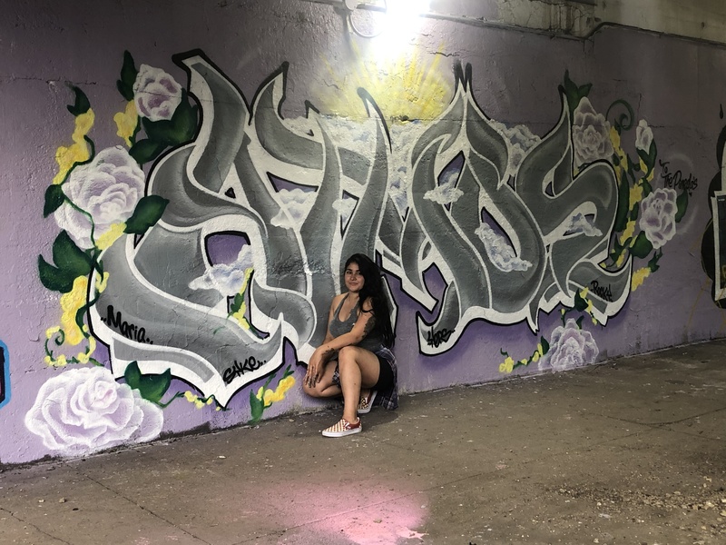  woman squats in front of outdoor spray painted mural. 