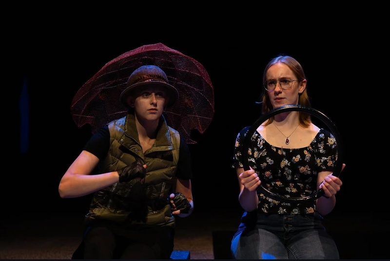  Two performers stand on stage. One is holding a black circle like a steering wheel while the other has a green tactical vest on. 