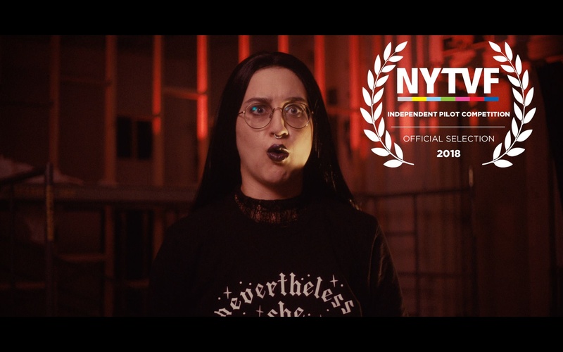  White, Queer, Non-binary Person in a long dark wig and glasses standing in front of a red wall. There is a NYTVF independent pilot competition stamp on the image. 