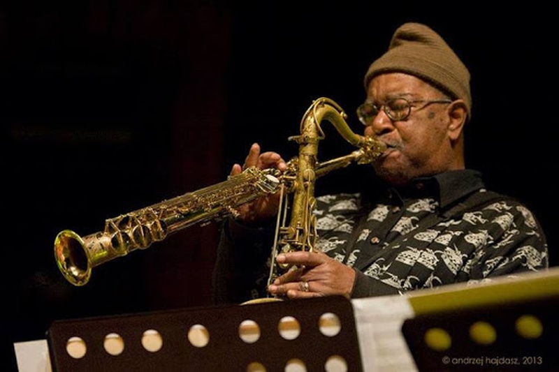  older black man with brown hat plays soprano and alto saxophone simultaneously 