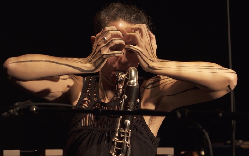  person holds their head while playing bass clarinet as light hits them from above 