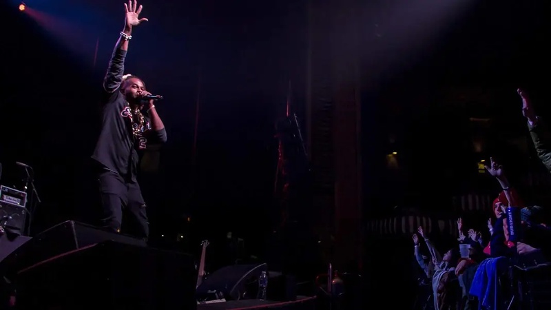  Black man points at the ceiling with light hitting him from above. crowd watches from the right. 