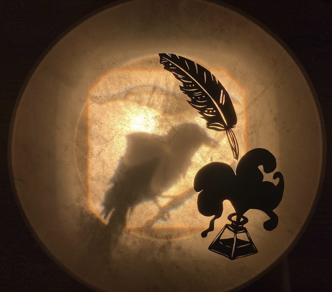  A shadow puppet of a bird, ink, and a quill cast in a warm toned light. 