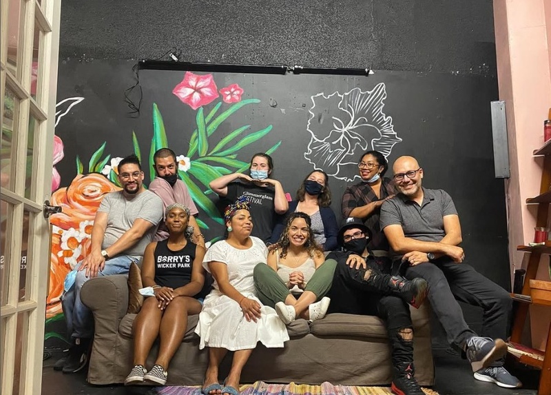  A group of 10 people all sit around a couch in difference poses. Some wear masks while others smile for the camera. Behind them is a black wall with a flowered mural. 