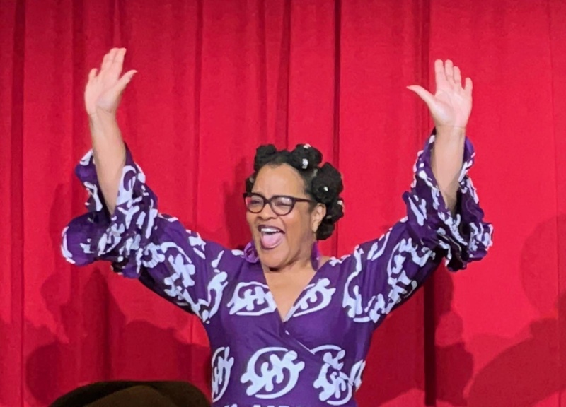  Black woman with hair in bantu knots wearing purple and white dress standing with arms raised in front of red background. 