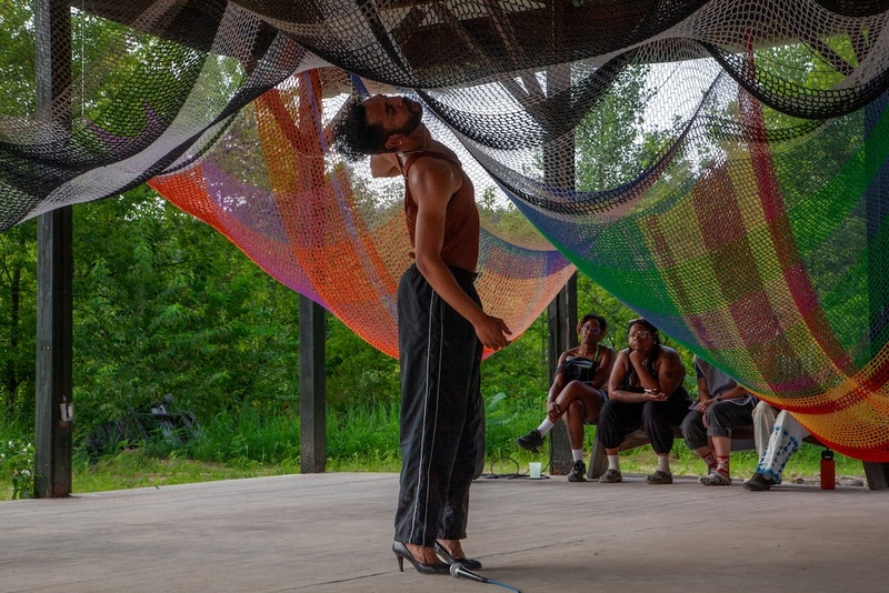  Light-skinned Black person with short black curly hair poses and they dance outside under a colorful netting woven from fabric. 