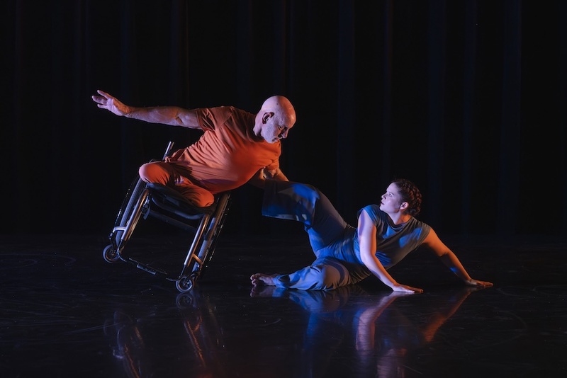  A white woman with red hair tied back and a man on a wheelchair perform in a dark room. The man is wearing orange and the woman is wearing blue. 