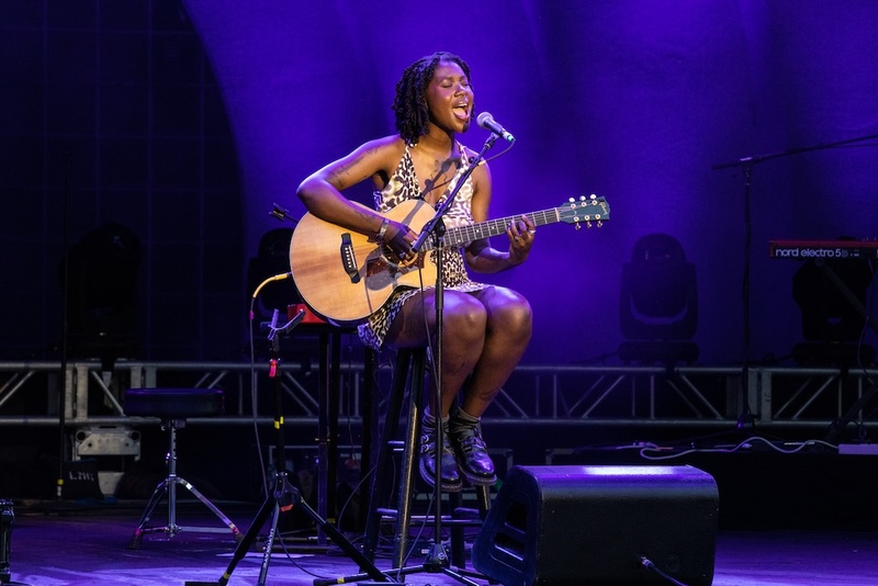  A Black girl with tattoos playing the guitar. She has short black locks and is wearing a black and white patterned dress. 