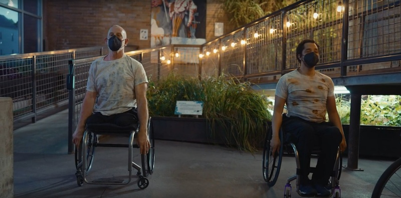  two people in wheelchairs and masks face the camera with lights behind them 