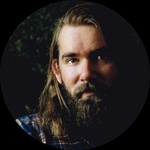 A headshot of a medium build white man with short, light-brown hair and a dark brown, medium length, full beard.