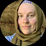 A headshot of a person smiling at the camera. They have light skin and blue/green eyes and are wearing a olive green hijab.