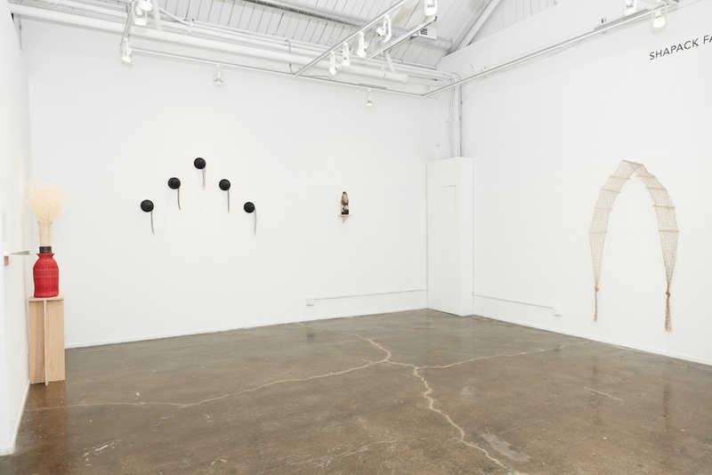  An installation shot in a white wall gallery. There are three sculptural pieces hanging on the wall and one red vase standing on a pedestal. 