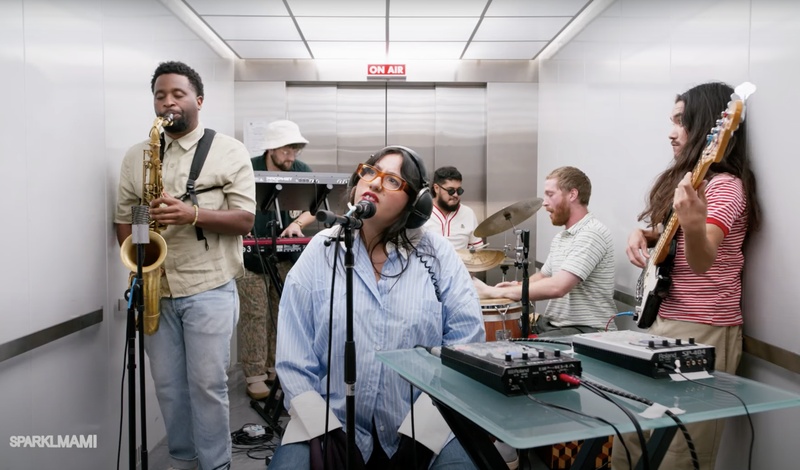  A woman with long brown hair and red glasses sits in front of a microphone. Behind her is a five piece band. They are in an elevator 