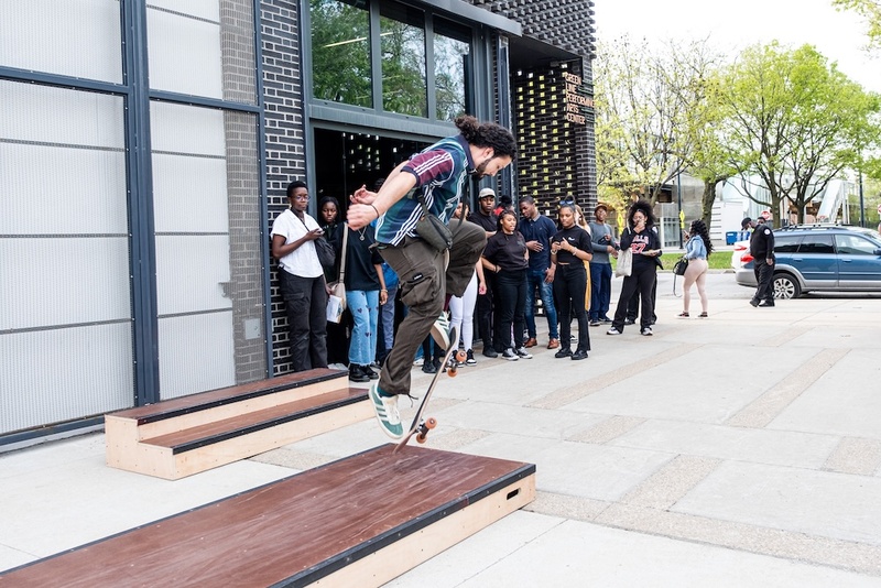  A teen does a trick on a skateboard outside a building with a crowd watching. 