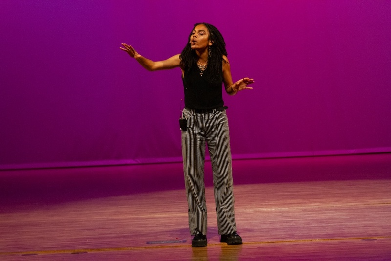  A Black, queer femme person with long black dreadlocks stands on a wooden floor with a purple wall behind them. They are performing poetry. 