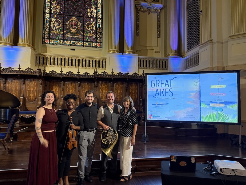  Five people stand on stage, smiling at the camera. Behind them is a stained glass window. To their right there is a projection on a screen titled The Great Lakes. 
