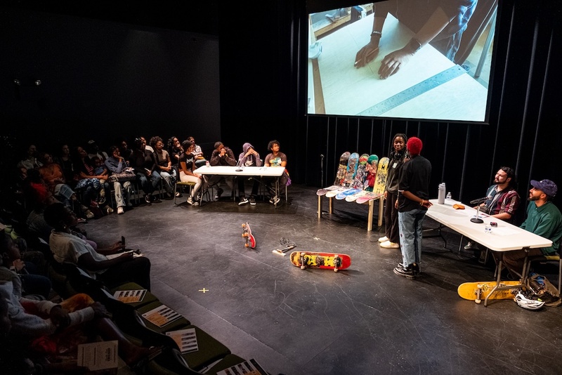  Two people stand on a stage filled with colorful skateboards. They are flanked by two tables with individuals sitting at them. Behind them is a video playing on a screen that shows an artist working on something on a drafting table. 