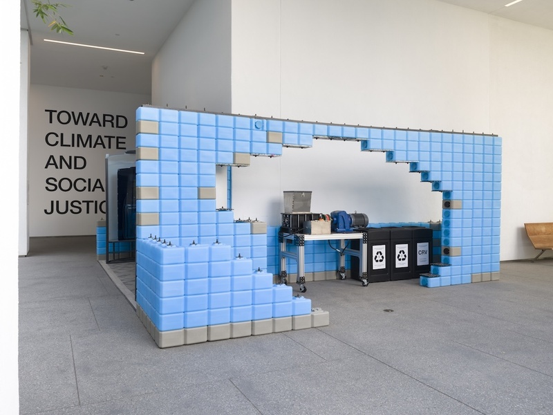  Blue blocks made up a wall inside a white walled gallery. The wall has holes in it where you can see the setup of recycling bins. 