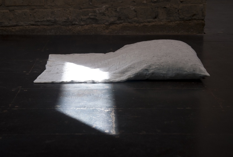  A white pillow lies on a dark, reflective tile floor. Sunlight streams through a skylight, casting angular light over part of the pillow. The background features a rough, light brown stone wall. The scene has a minimalist and serene atmosphere. 