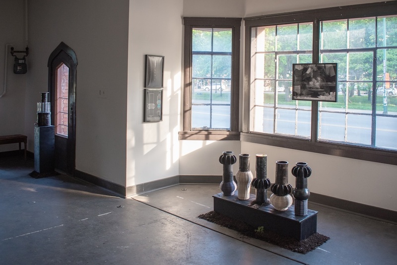  A photo of an installation inside a gallery. There is a sculpture of different sized vases sitting on a pedestal in front of a window. 