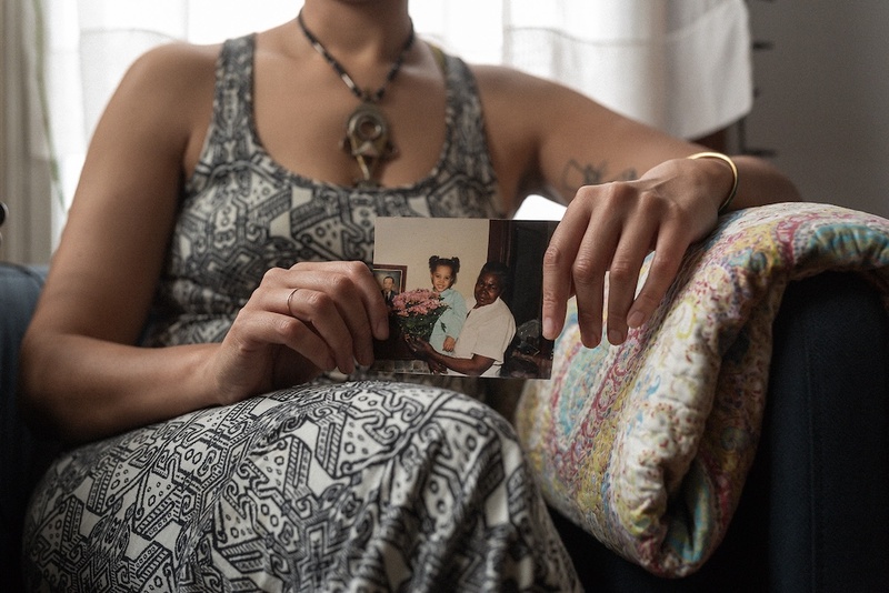  A figure sits on a chair, only visible from the shoulders down. They are wearing a black and white patterned dress, a necklace with a large pendant and are holding a photograph of a young child with an older figure holding flowers. 
