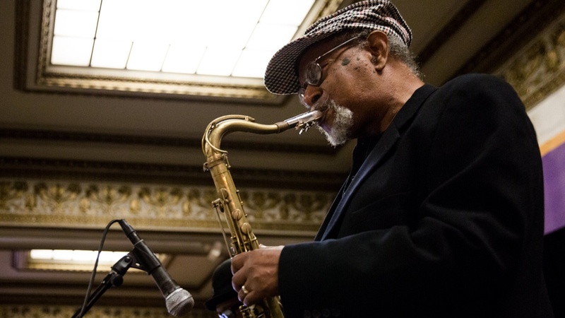  older black man wearing checkered paperboy cap plays saxophone into a microphone 