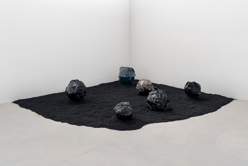  Image of a sculpture in a white wall gallery. In the corner there is black sand that extends out. On top of the sand there are ceramic orbs with textured surfaces. 
