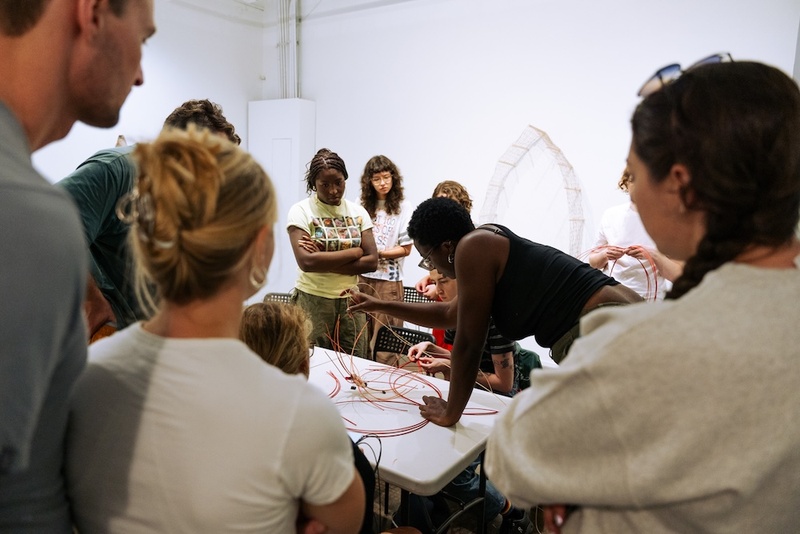  A black woman with glasses, queer presenting with short black hair leads a basket weaving workshop indoors. 