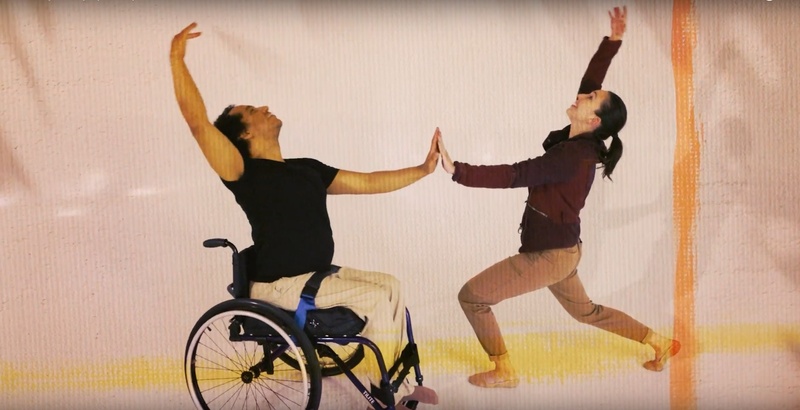  one dancer in wheelchair and one standing dancer touch hands and hold pose in beige room 